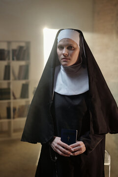 Portrait of young Caucasian woman dressed as nun standing in prison library holding small book with cross on cover looking with serious expression