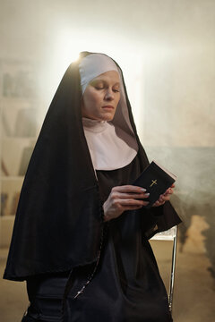 Caucasian woman dressed as nun sitting in prison library holding closed book with cross on cover, eyes closed, appearing to pray or meditate in quiet moment