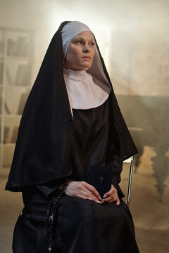 Caucasian woman dressed as nun sitting in prison library holding closed book with cross on cover, looking away with serious expression, rosary beads in hand