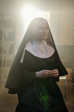 Caucasian woman dressed as nun sitting in prison library holding closed book with cross on cover, looking forward with calm expression, sunlight streaming behind