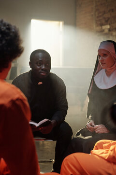 Black man wearing clerical collar reading book while sitting with Caucasian woman dressed as nun and group of prisoners in orange uniforms inside prison library