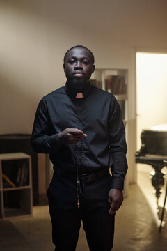 Portrait of young adult Black priest standing in prison library holding rosary beads, wearing clerical collar, looking directly at camera with serious expression, bookshelves in background