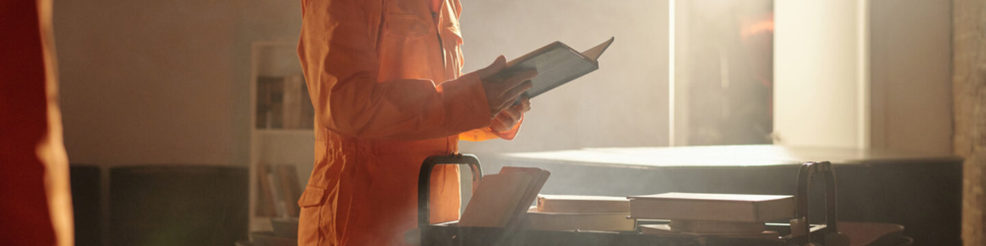Man standing in prison library holding open book while selecting reading material from book cart with several stacked books visible in correctional facility setting