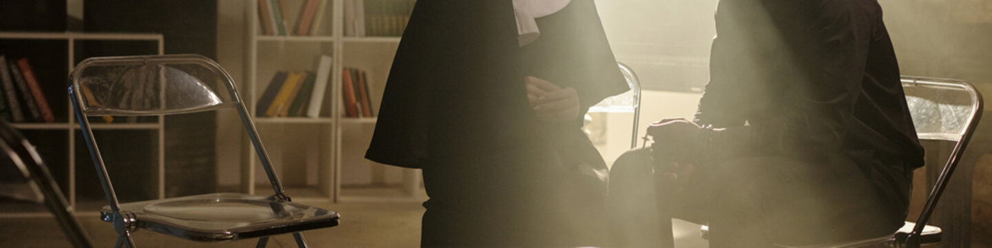 Nun sitting on chair holding hands together while young adult Black priest sitting opposite holding book with cross in prison library setting