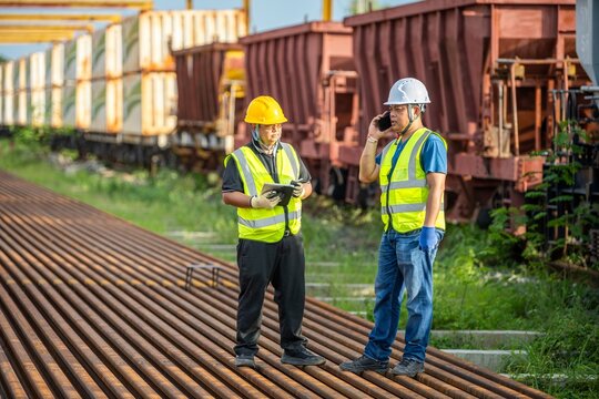 Communication in Industry: Engineer Talking on Phone, Another with Tablet at Railway Site Outdoor Engineering: Two Confident Engineers Standing and Planning Maintenance on Iron Train Tracks.