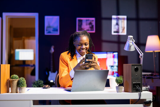 Happy woman at home having fun looking at social media reels done by online content creators. Smiling african american teenager relaxing by using mobile social network app at laptop desk