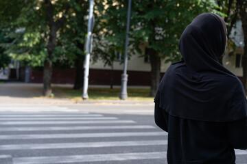 Woman in black attire stands at crosswalk observing surroundings in a park area on a sunny day