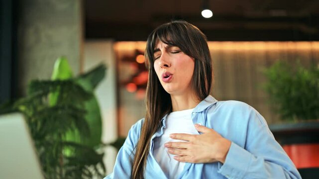 Distressed woman in blue shirt, white t-shirt experiences sudden chest pain, anxiety. She clutches chest, showing discomfort while working on laptop in modern office. Depicts health issues and stress.