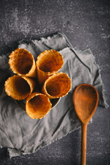 Five waffle cones are neatly arranged in a bowl on a gray textured cloth, with a wooden spoon...