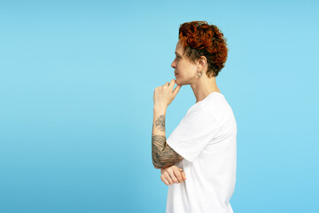 Portrait of woman with short red hair and tattoos, wearing white t shirt against blue background