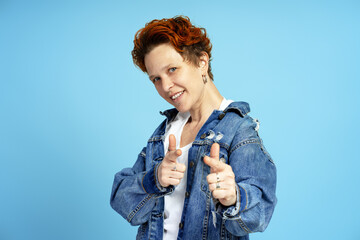 Portrait of joyful young woman with red hair and piercings wearing denim jacket on blue background