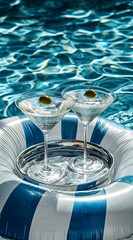 Two elegant martini glasses sit on a striped pool float, with clear water shimmering around them, creating a refreshing and inviting summer vibe perfect for relaxation.