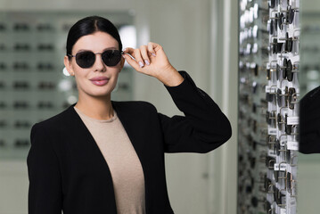 Attractive Caucasian young brunette woman in black suit trying on and choosing sunglasses to buy at...