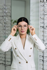 Attractive Caucasian young brunette woman in white suit trying on and choosing eyeglasses for buying at an optical store. Lifestyle, optometry, eyesight, assortment of glasses
