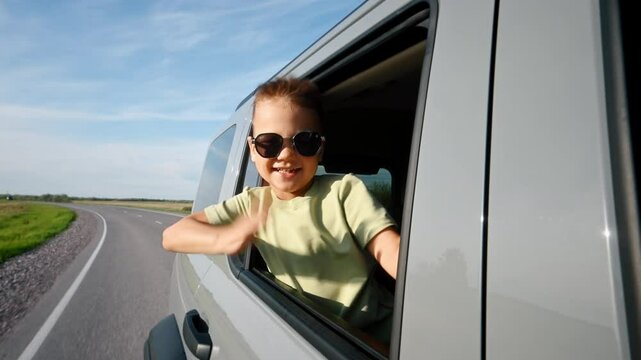 A little boy going on summer vacation on sunny day, looking out of the car window and waving hand happily, joy of family travel, childhood and carefree. Road going through endless fields