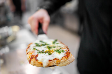 Close-up of a hot pizza slice with melted cheese, tomato sauce, and herbs on a metal spatula, held...