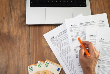 Fototapeta premium Man points with pen at Employee Withholding Certificate tax form examining steps of filling blank boxes properly at workplace with euro banknotes and laptop