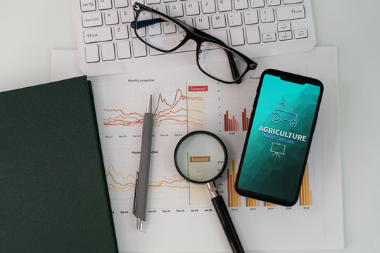 A smartphone displaying Agriculture Market Outlook report is placed atop a stack of papers with graphs and charts, accompanied by yellow notebook, keyboard, glasses and magnifying glass 