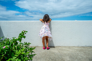 A young Japanese girl wearing a pink butterfly dress and sandals leans over a white wall to look at...