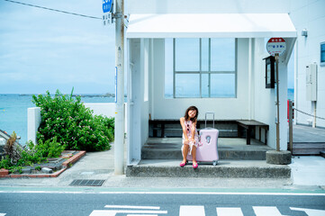 Young Japanese girl sitting happily at a white bus stop near the ocean with a pink suitcase,...