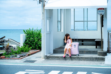 Japanese girl in a butterfly print dress sitting on a pink suitcase at a white seaside bus stop,...