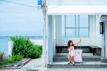 Young Japanese girl with long dark hair sitting on a suitcase at a seaside bus stop, enjoying...