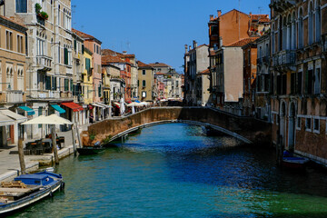 Lively Venetian canal with arched pedestrian bridge, colorful facades, waterfront cafés and boats under a bright blue sky – authentic travel scene showcasing everyday charm of Venice’s city life