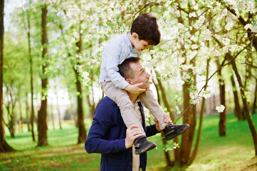 father and son enjoying spring nature in blooming park. child on father's shoulders, surrounded by vibrant green trees and white blossoms. perfect family bonding moment outdoors.