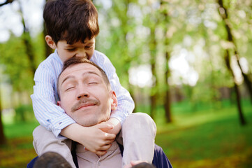 father carrying son on shoulders in sunny park, both smiling, embracing bond and joyful connection....