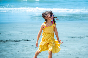 Young Japanese girl in a yellow dress and sunglasses crouching playfully on a sandy beach, enjoying...