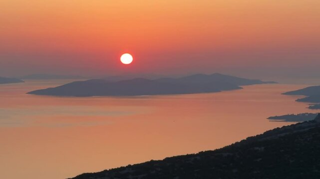 As the red sun descends below the horizon, its glow illuminates the sky in shades of orange and pink. The silhouettes of islands and rugged mountains create a breathtaking foreground against the majes