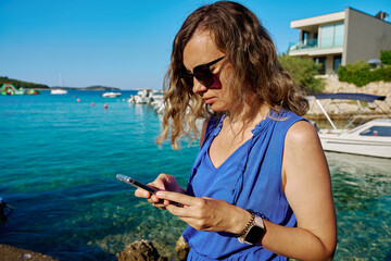 Woman in sunglasses and blue dress using smartphone by sea on sunny day. Female tourist texting by...