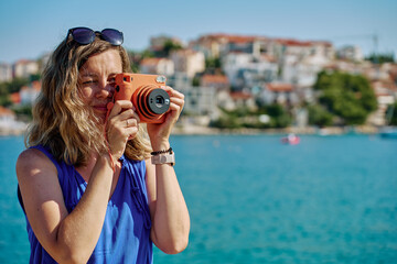 Woman standing near sea holding instant camera and taking photo. Female tourist at coastal resort...
