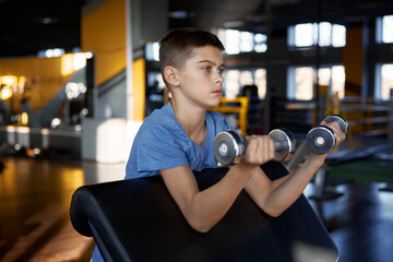 Teenage boy pumping biceps enjoying training with dumbbells at gym