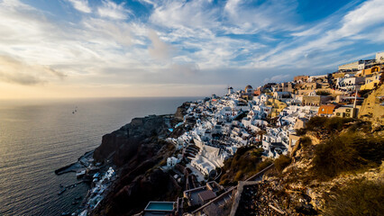 The iconic view of Oia glows warmly during the golden hour, as soft sunlight bathes the whitewashed buildings and blue domes. The vibrant colors and glowing caldera create a radiant atmosphere