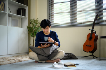 Young man sitting cross egged on the floor, drinking coffee and browsing tablet at home