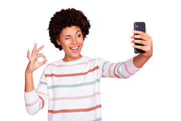 Photo of pretty dark skin lady holding telephone making selfies showing okey symbol winking eye wear striped pullover isolated pastel pink color background
