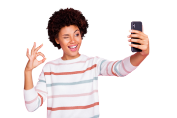 Photo of pretty dark skin lady holding telephone making selfies showing okey symbol winking eye wear striped pullover isolated pastel pink color background