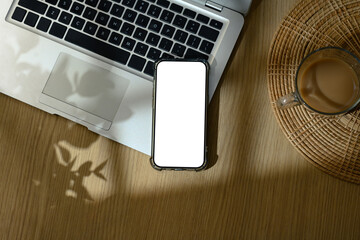 Smartphone with blank white screen placed on laptop keyboard next to coffee cup on wooden desk