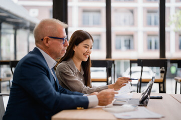 Two businesspeople collaborating on a project, analyzing financial charts and data on a laptop while reviewing documents at a shared table in a modern office