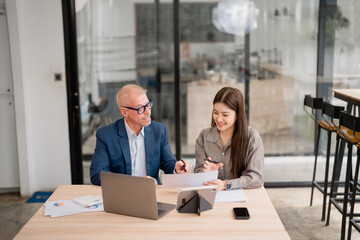 Businessman and businesswoman sitting at desk using laptop, tablet and documents, discussing about...