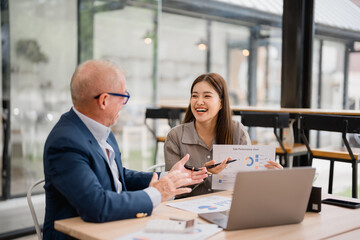 Senior businessman and young businesswoman laughing and discussing a sales performance chart during a productive meeting in a modern office filled with natural light