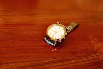 A gold watch with a black band sits on a wooden table. The watch is set to the time of 10:30