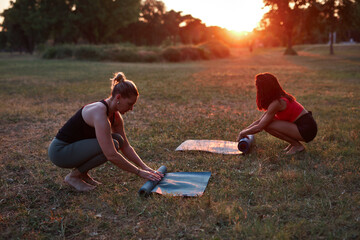 Two women folding and unfolding yoga mat in the park during sunset sunrise time.