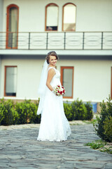 A bride is standing in front of a building with a bouquet of flowers in her hand. She is wearing a...