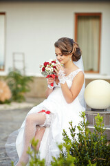 A bride is sitting on a stone wall, holding a bouquet of flowers. She is wearing a white dress and...