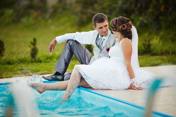 A bride and groom are sitting in a pool, with the bride wearing a white dress. The man is wearing a...