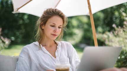Young woman working outdoors with laptop under umbrella in garden, enjoying remote work, digital lifestyle, summer weather and iced coffee relaxation.