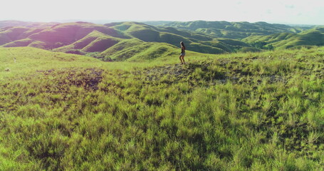 Aerial view capturing a young woman relishing her trekking adventure while walking across the lush grassy hills of Sumba Island, Indonesia, during a breathtaking sunset