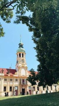 Prague Loreta. Historic building with clock tower surrounded by lush greenery and blue sky, showcasing architectural beauty and vibrant colors in a serene outdoor setting